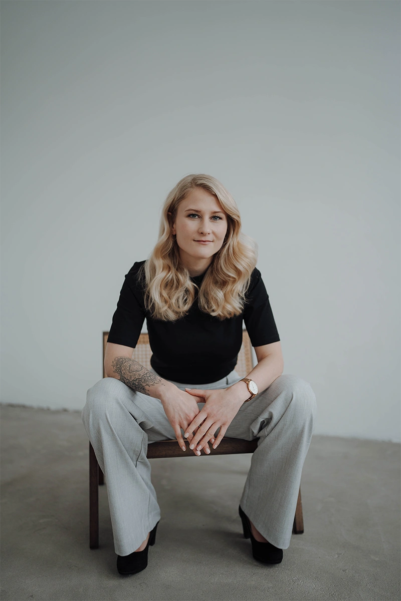 Anja Götzfried, Webdesignerin, sitzt mit langen gewellten Haaren auf einem Stuhl und trägt schwarzen Oberteil, graue Hose und Armbanduhr
